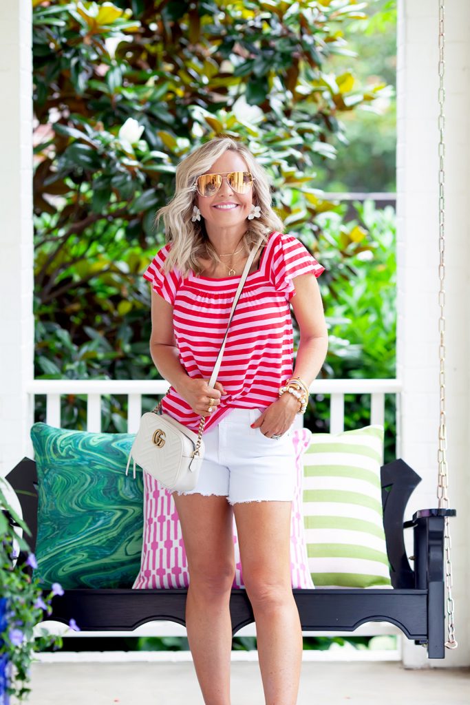 Gibson x Hi Sugarplum Summer of Color Collection by top us fashion blog, Hello Happiness: image of standing woman outside in front of a porch swing and wearing a Malta Knit Square Neck Ruffle Sleeve Tee in Red and Pink Stripe, Gemma shorts, Espadrilles, Maramont Crossbody, and x Desi Perkins High Key 62mm. 