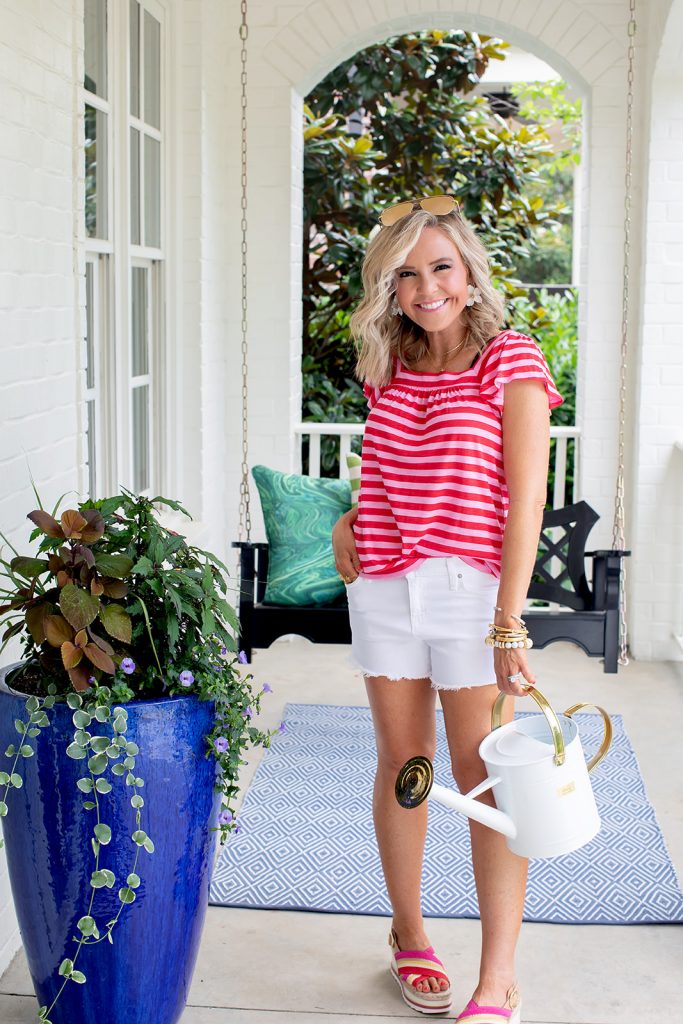 Gibson x Hi Sugarplum Summer of Color Collection by top us fashion blog, Hello Happiness: image of woman outside holding a white watering can and wearing a Malta Knit Square Neck Ruffle Sleeve Tee in Red and Pink Stripe, Gemma shorts, Espadrilles, Maramont Crossbody, and x Desi Perkins High Key 62mm. 