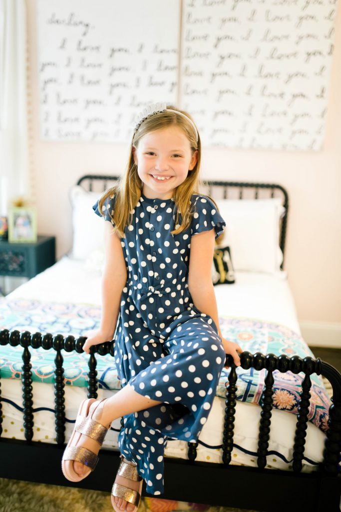 Fashionistas in Training + Nordstrom Kids Clothing by popular Nashville life and style blog, Hello Happiness: image of sitting on the edge of her bed and wearing a Nordstrom Tucker + Tate Wide Leg Romper, Maniere Metallic Headband, and Mel by Melissa Cosmic Sandal. 