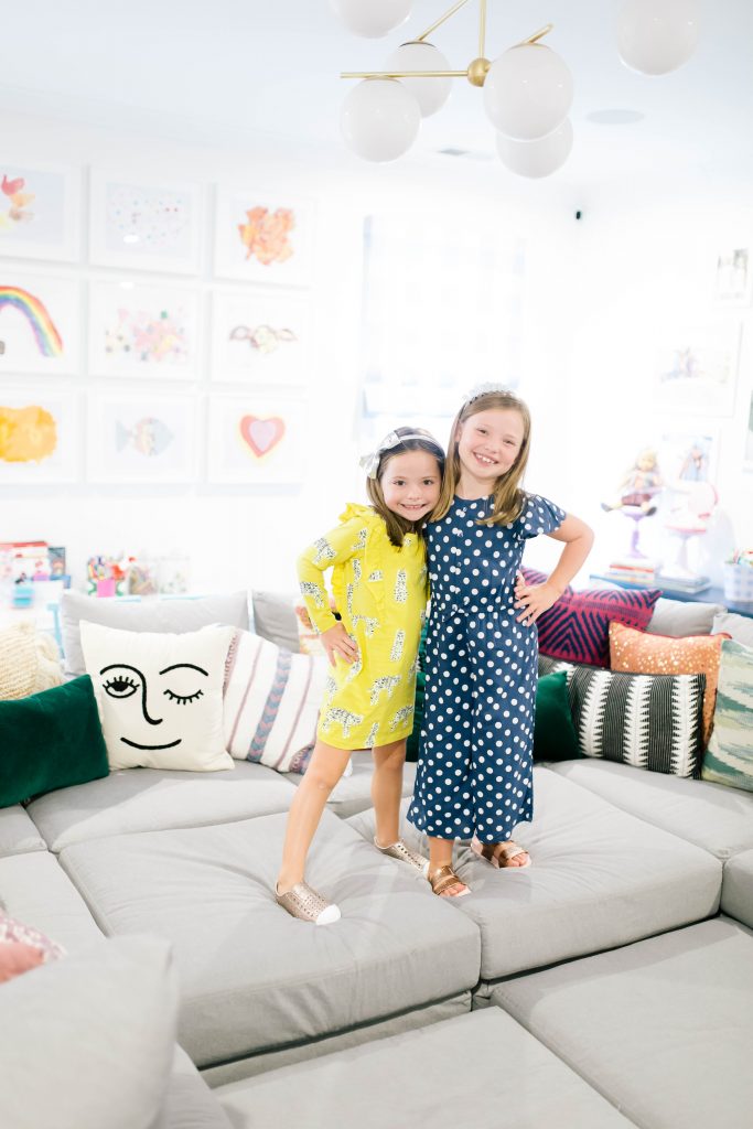Fashionistas in Training + Nordstrom Kids Clothing by popular Nashville life and style blog, Hello Happiness: image of two girls standing on a sectional couch and wearing a Nordstrom Tea Collection Ruffle Dress, Native Shoes Jefferson Bling Glitter Slip-On Vegan Sneaker, Tucker + Tate Wide Leg Romper, Maniere Metallic Headband, and Mel by Melissa Cosmic Sandal. 