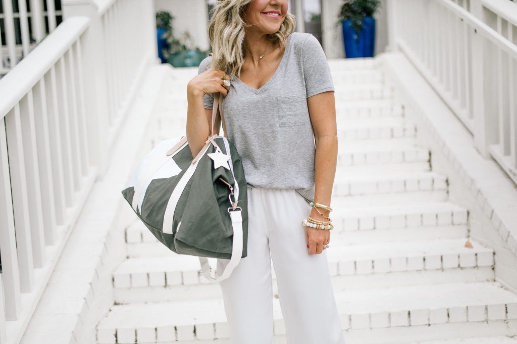 We Dress America + Walmart Travel Style for a Successful Getaway by top US fashion blog, Hello Happiness: image of woman standing outside on white brick steps wearing a gray v-neck pocket t-shirt, sunglasses, white pants, and holding a large gray duffle bag with a white star print and white straps.