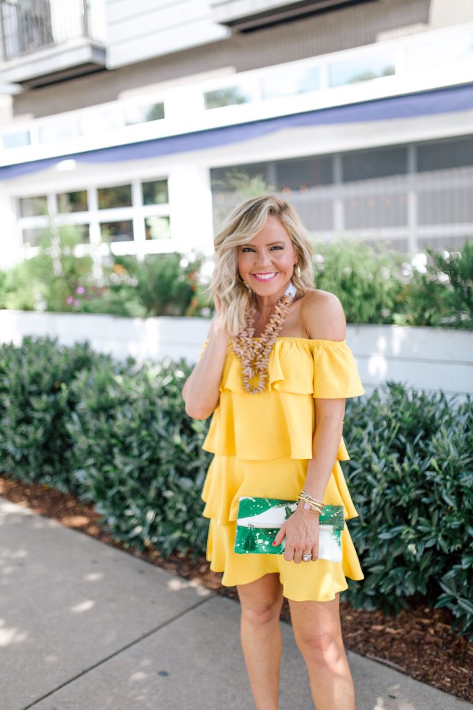 Those Hot Summer Nights... A New Collection from Gibson! by popular Nashville fashion blog Hello Happiness: image of a woman standing outside on a sidewalk next to a white building wearing a yellow Gibson Hot Summer Nights Natalie Off The Shoulder Ruffle Dress, Hearne Dry Goods Co. Tahiti Beach necklace, Steve Madden Bree Leopard wedge sandals, and holding a H. Howorth Art Emerald Green Clutch.