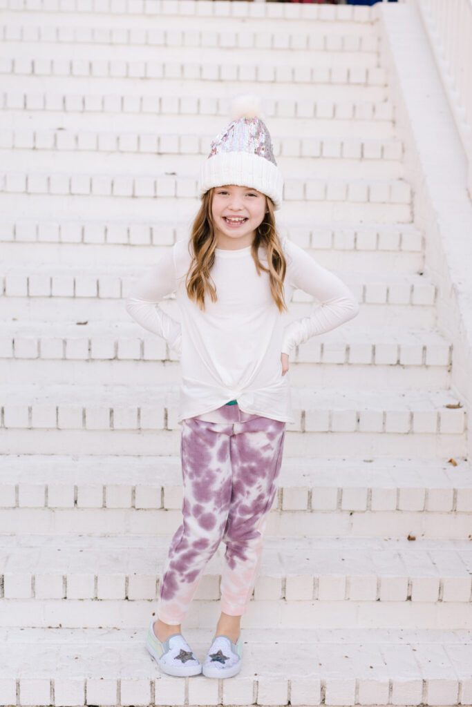 Nordstrom Fashion by popular Nashville fashion blog, Hello Happiness: image of a girl sitting on a white brick wall and wearing a Tucker and Tate sequin pom beanie, Nordstrom Zella Kids' Tie Dye Joggers, and Zella Kids' Peaceful Twist Front T-Shirt.