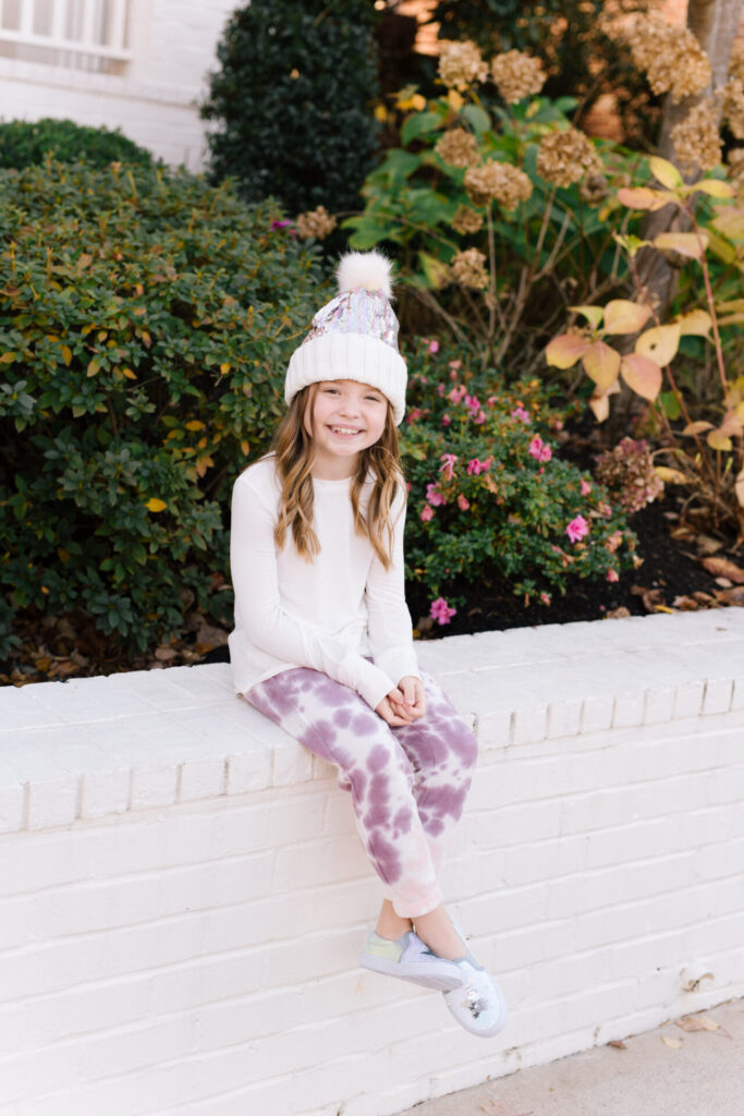 Nordstrom Fashion by popular Nashville fashion blog, Hello Happiness: image of a girl sitting on a white brick wall and wearing a Tucker and Tate sequin pom beanie, Nordstrom Zella Kids' Tie Dye Joggers, and Zella Kids' Peaceful Twist Front T-Shirt.