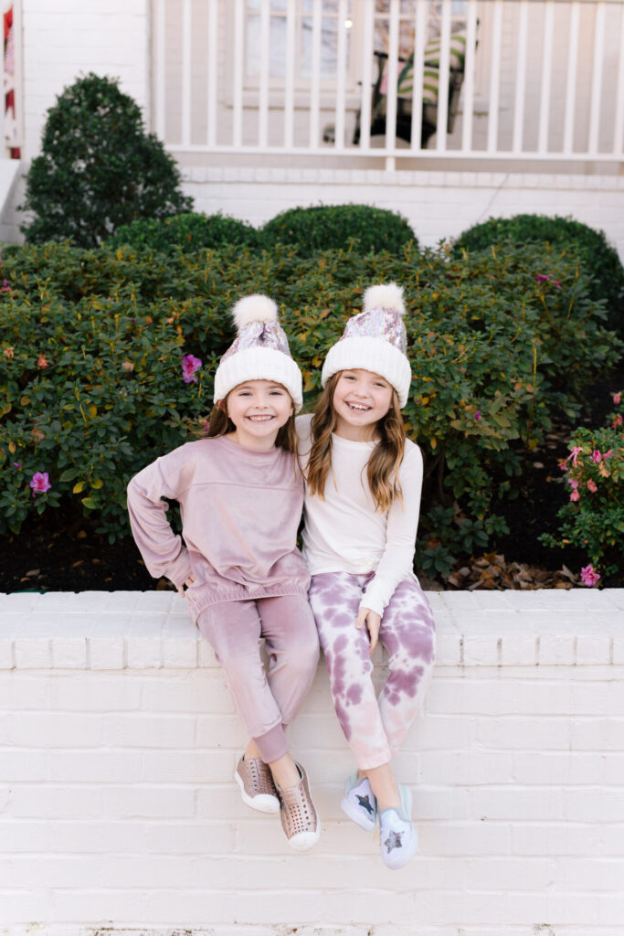 Nordstrom Fashion by popular Nashville fashion blog, Hello Happiness: image of two girls sitting on a white brick wall and wearing a Tucker and Tate sequin pom beanie, Nordstrom Zella Girl Kids' Cozy Active Pullover, Nordstrom Zella Girl Kids' Velour Cozy Active Joggers, Zella Girl Kids' Tie Dye Joggers, and Zella Girl Kids' Peaceful Twist Front T-Shirt.