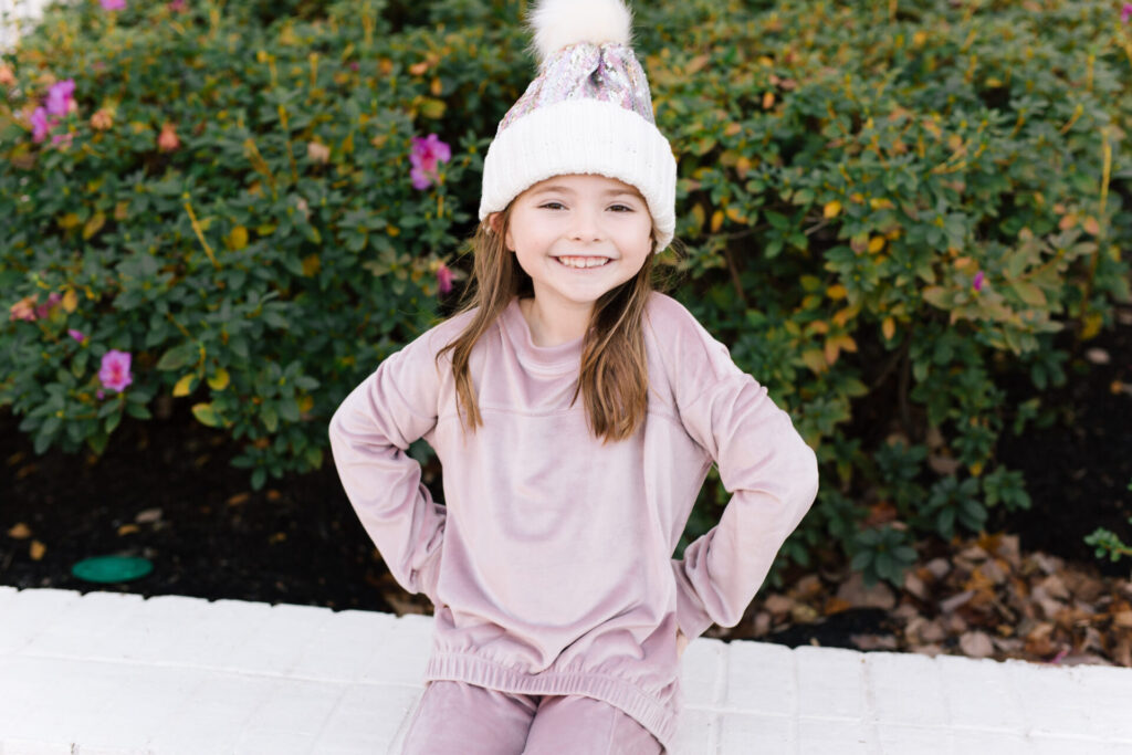 Nordstrom Fashion by popular Nashville fashion blog, Hello Happiness: image of a girl sitting on a white brick wall and wearing a Tucker and Tate sequin pom beanie, Nordstrom Zella Girl Kids' Cozy Active Pullover, and Nordstrom Zella Girl Kids' Velour Cozy Active Joggers.