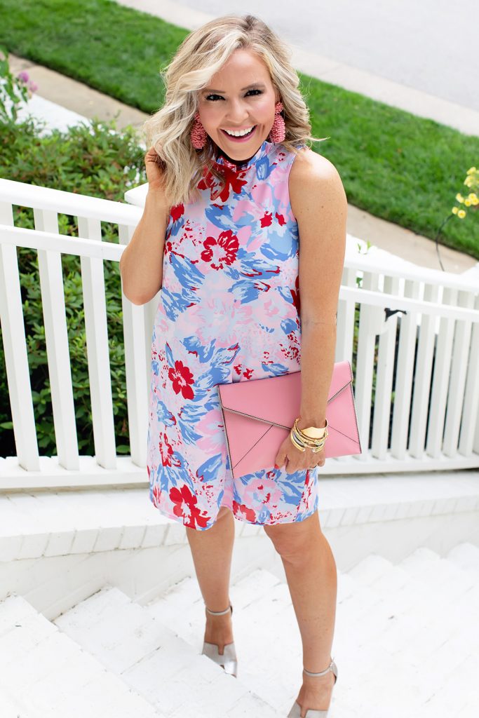 Gibson x Hi Sugarplum Summer of Color Collection by top us fashion blog, Hello Happiness: image of smiling woman sitting outside by a white flowering bush and wearing the Sardinia Ruffle Neck Date Dress in Vista Cherry, gold wedges, beaded earrings, and holding the pink Leo clutch.