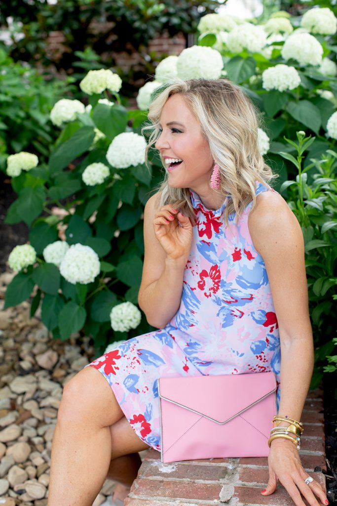 Gibson x Hi Sugarplum Summer of Color Collection by top us fashion blog, Hello Happiness: image of smiling woman sitting outside by a white flowering bush and wearing the Sardinia Ruffle Neck Date Dress in Vista Cherry, gold wedges, beaded earrings, and holding the pink Leo clutch.