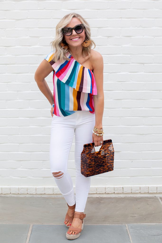 Gibson x Hi Sugarplum Summer of Color Collection by top us fashion blog, Hello Happiness: image of smiling woman standing outside and wearing an Ibiza One Shoulder Ruffle Top in Summer Stripe, bamboo earrings, air heart sunglasses, ripped skinny jeans, Audrina sandals, and acrylic bag. 