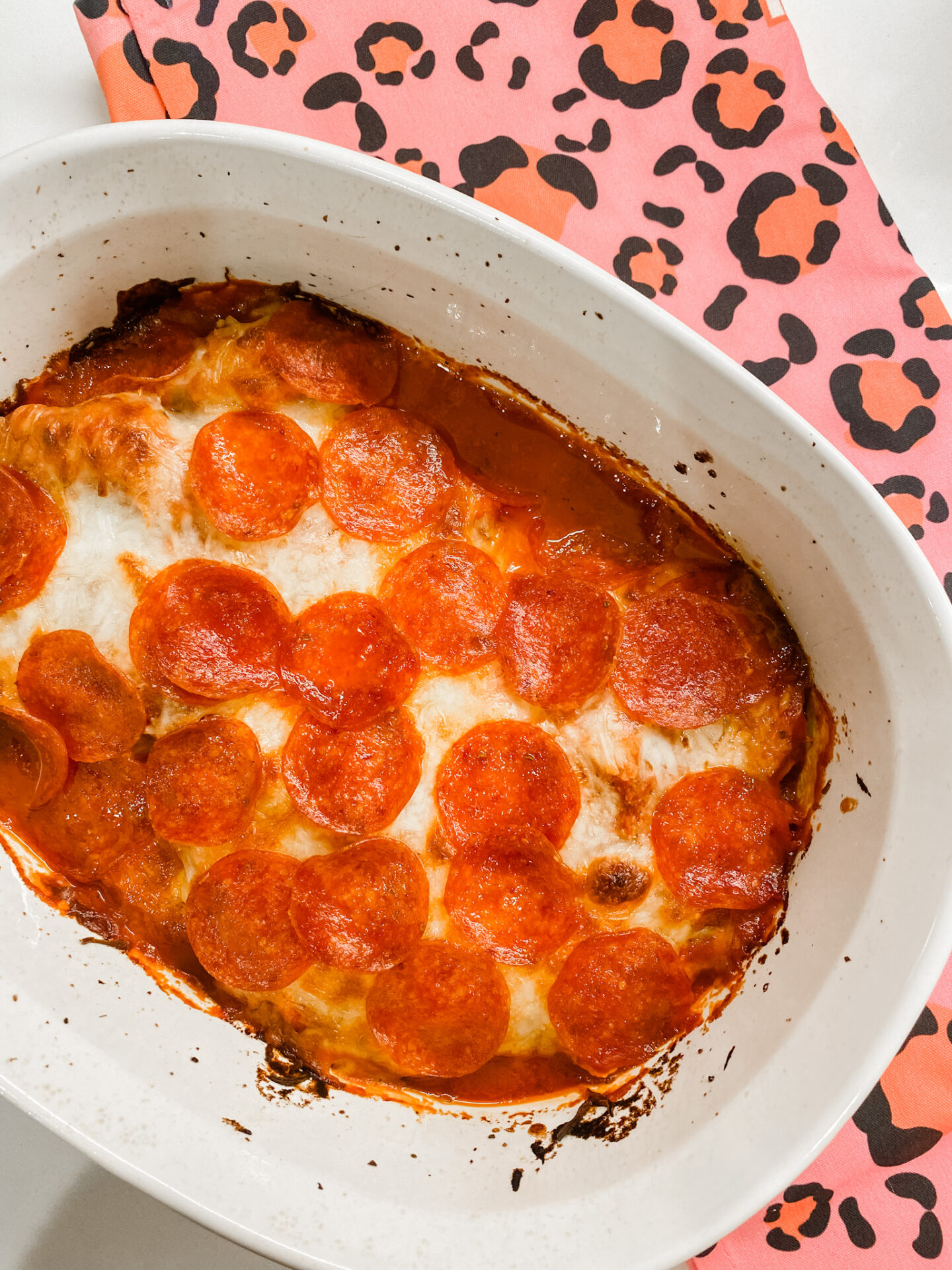 Pepperoni Pizza Chicken Bake by popular Nashville lifestyle blog, Hello Happiness: image of a pepperoni pizza chicken back in a white ceramic dish next to a pink and leopard print towel. 