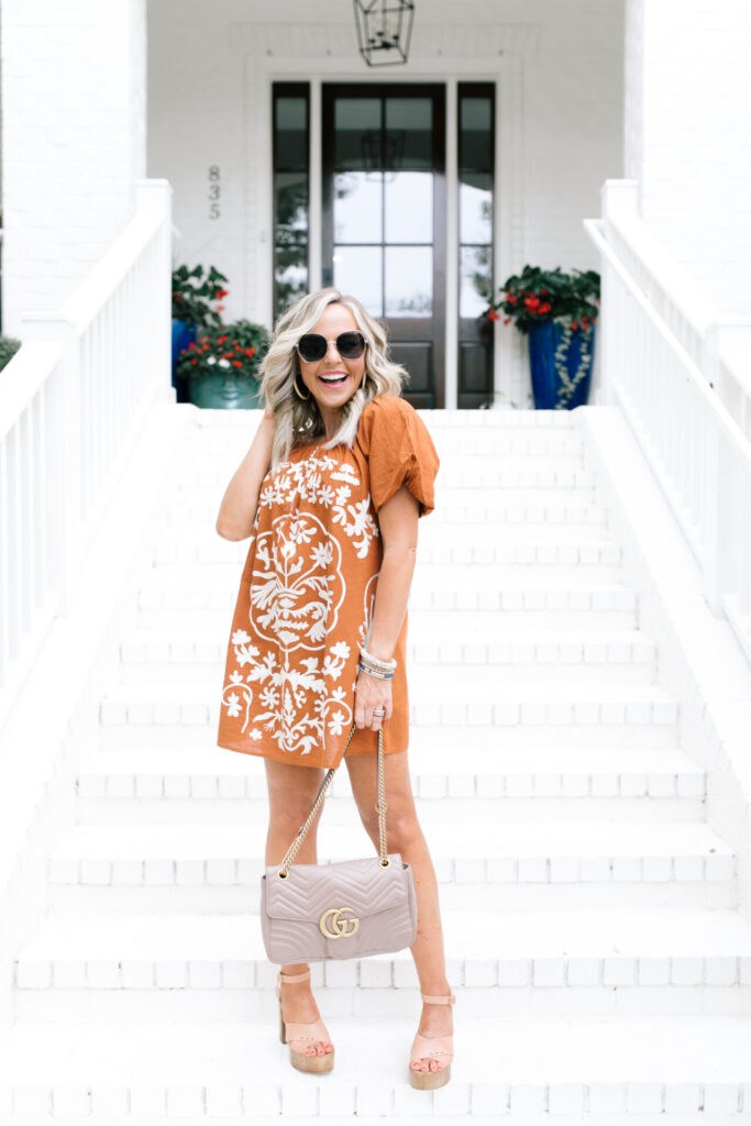 Rust Color by popular Nashville fashion blog, Hello Happiness: image of Natasha Stoneking standing on her white brick steps and wearing a Free People Free People Fiona Embroidered Mini Dress, Quay Big Love sunglasses, Julie Vos Barcelona Statement Ring, and holding a Gucci bag. 