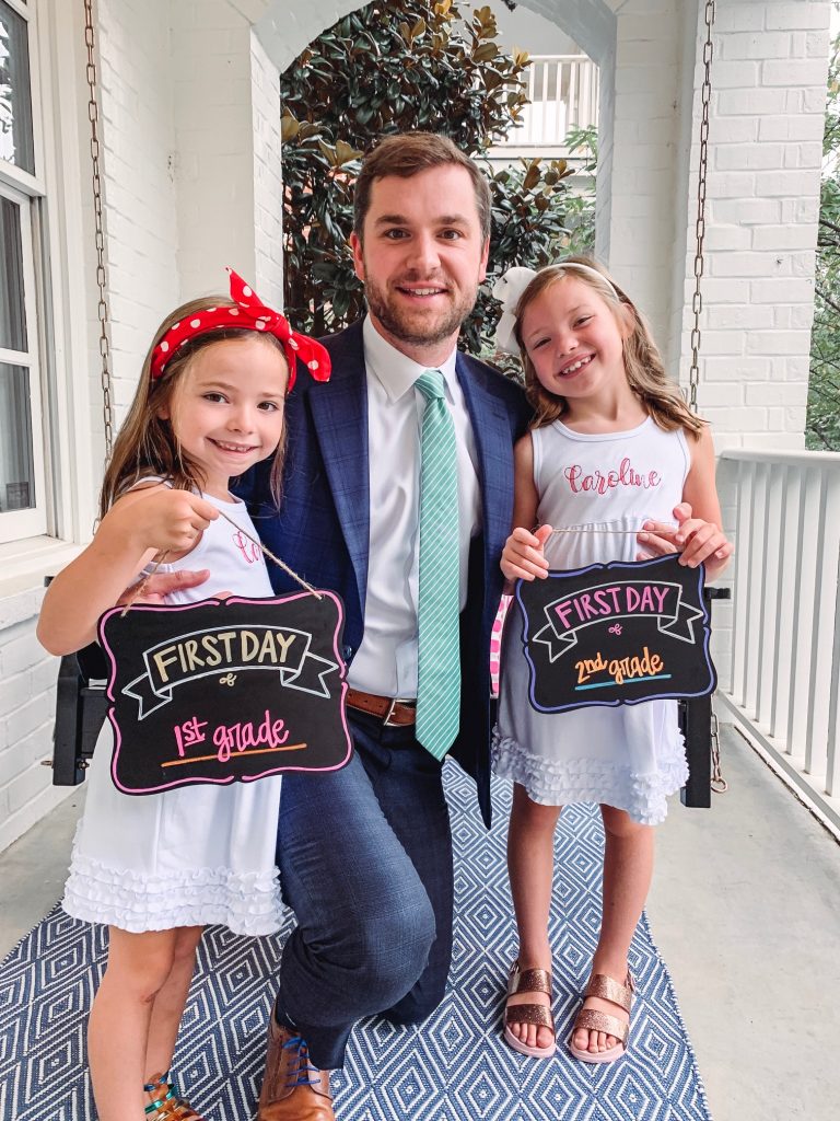 Caroline and Carson Go Back to School... And They're Off! by popular Nashville blog, Hello Happiness: image of two little girls standing outside with their dad and wearing an Elegant Goods Shop dress and holding a Lovely Little Designs chalkboard sign. 