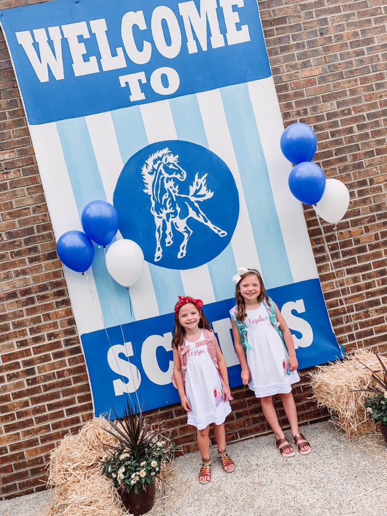 Caroline and Carson Go Back to School... And They're Off! by popular Nashville blog, Hello Happiness: image of two little girls standing outside in front of a 'Welcome Back to School' sign and wearing an Elegant Goods Shop dress.
