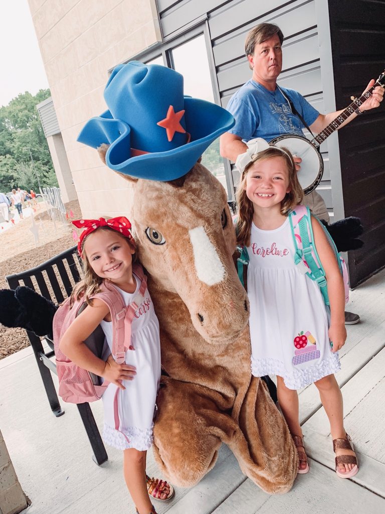 Caroline and Carson Go Back to School... And They're Off! by popular Nashville blog, Hello Happiness: image of two little girls standing outside with a horse mascot and wearing an Elegant Goods Shop dress.