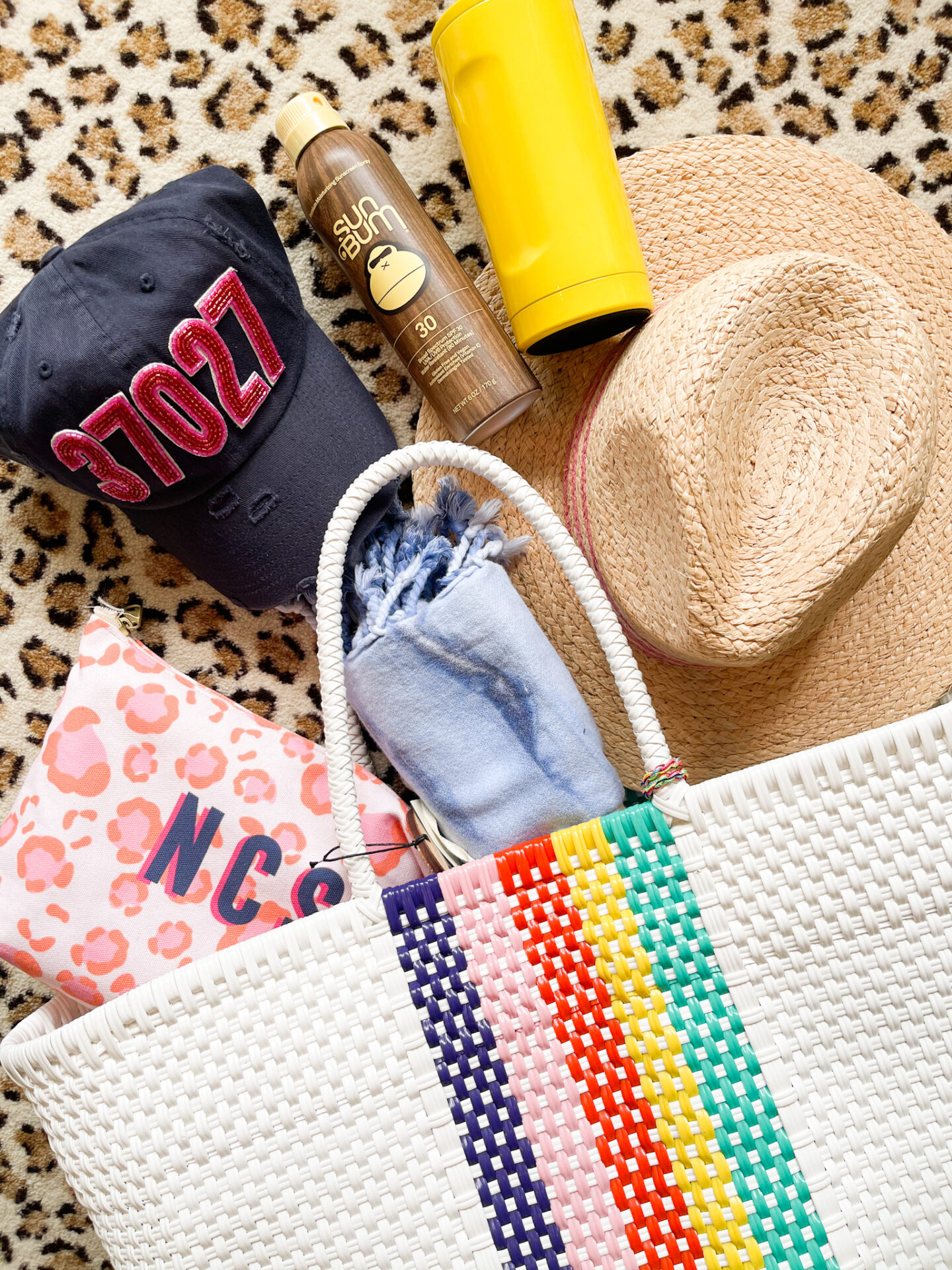 Spring Break Outfits by popular Nashville fashion blog, Hello Happiness: image of a white woven tote bag next to a straw sun hat, blue baseball cap, sun Bum sunscreen, and a personalize cosmetic tote. 