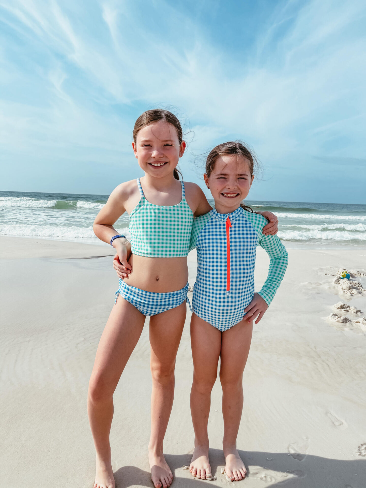 Spring Break Outfits by popular Nashville fashion blog, Hello Happiness: image of two young girls standing together on the beach and wearing a gingham tankini set and gingham rashguard. 