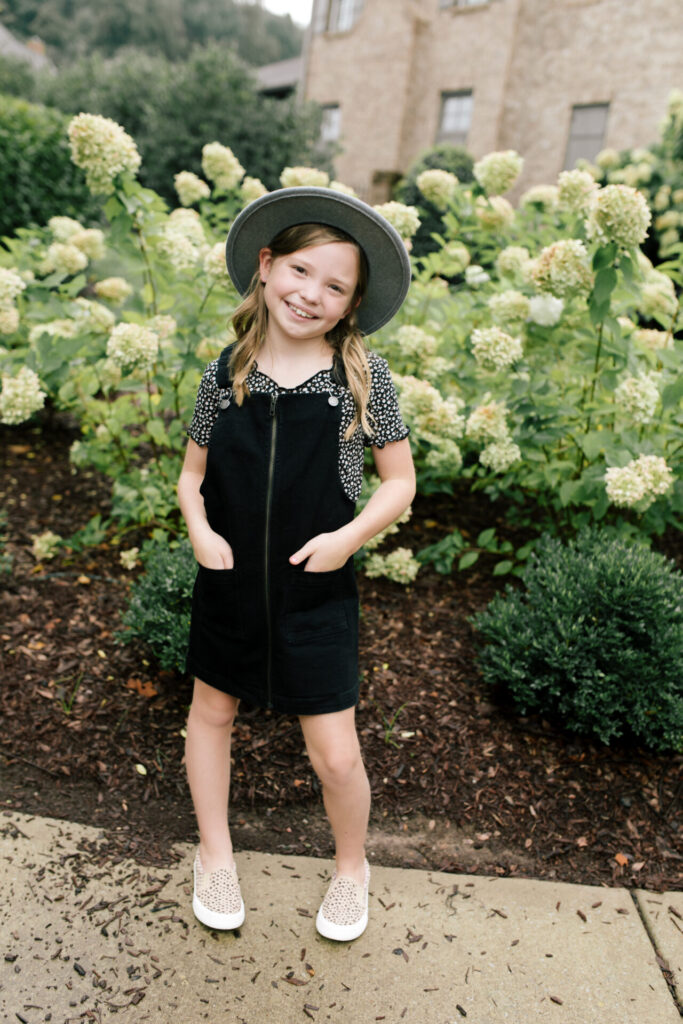 Tween Fashion by popular Nashville fashion blog, Hello Happiness: image of a girl standing outside in front a bush with white flowers and wearing a Target Art Class black pinafore dress, Target thermal floral tee, Target studded sneakers, and Target grey felt floppy studded fedora.