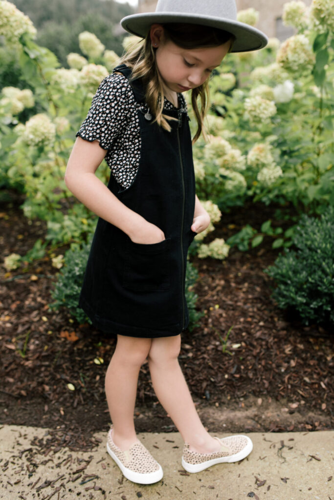 Tween Fashion by popular Nashville fashion blog, Hello Happiness: image of a girl standing outside in front a bush with white flowers and wearing a Target Art Class black pinafore dress, Target thermal floral tee, Target studded sneakers, and Target grey felt floppy studded fedora.