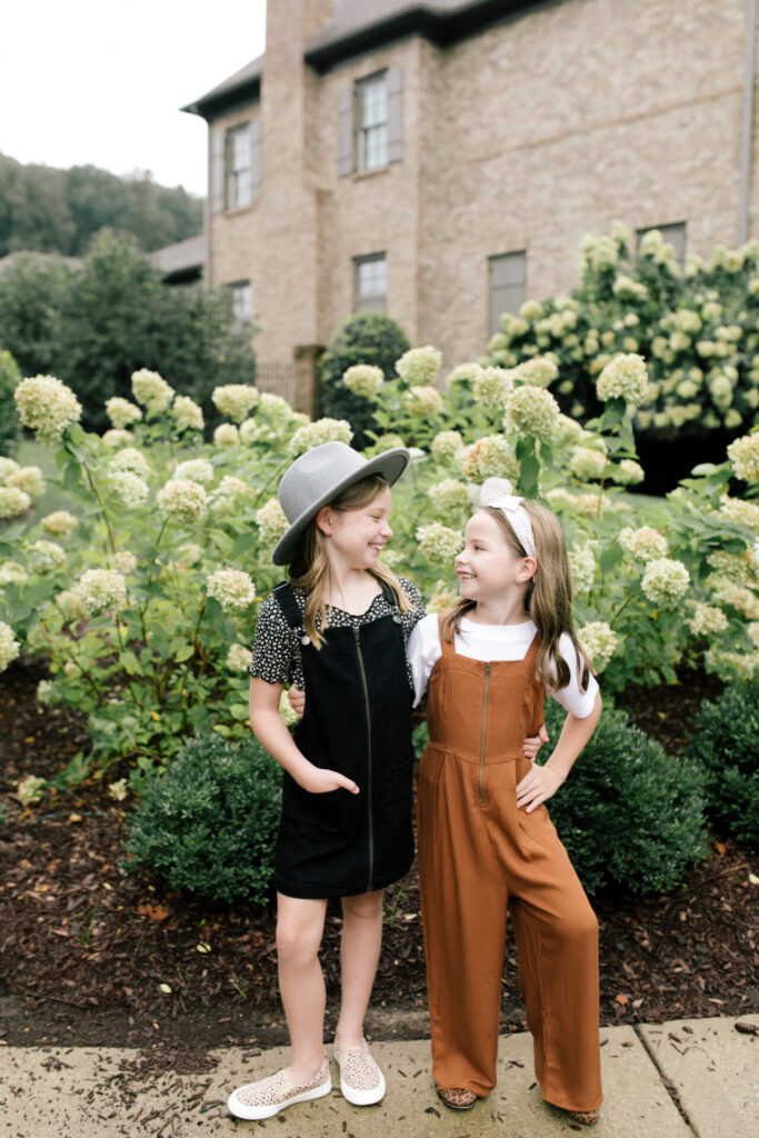 Tween Fashion by popular Nashville fashion blog, Hello Happiness: image of two girls standing outside together in front a bush with white flowers and wearing a Target Art Class black pinafore dress, Target thermal floral tee, Target studded sneakers, Target grey felt fedora, Target zip front utility jumpsuit, Target classic boxy t-shirt, and Target leopard print booties. 