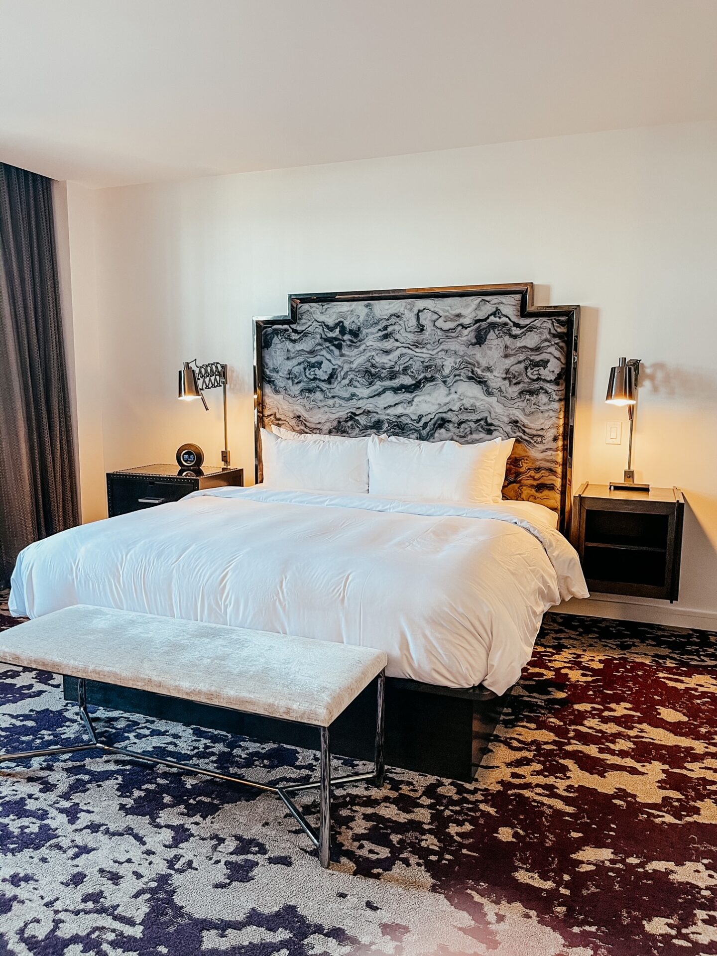 Girls Getaway by popular Nashville travel blog, Hello Happiness: image of a king size bed with white bedding and a marble headboard in a room with grey drapes and purple and white carpet. 