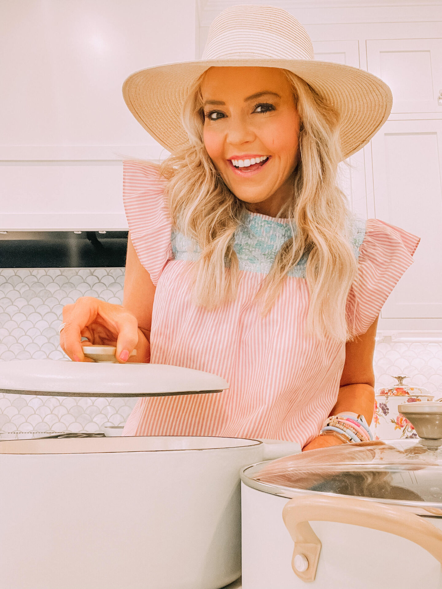 Beautiful by Drew Barrymore by popular Nashville life and style blog, Hello Happiness: image of a woman wearing a straw hat and orange and white stripe top standing next to a white cooking pot from the Beautiful by Drew Barrymore collection. 
