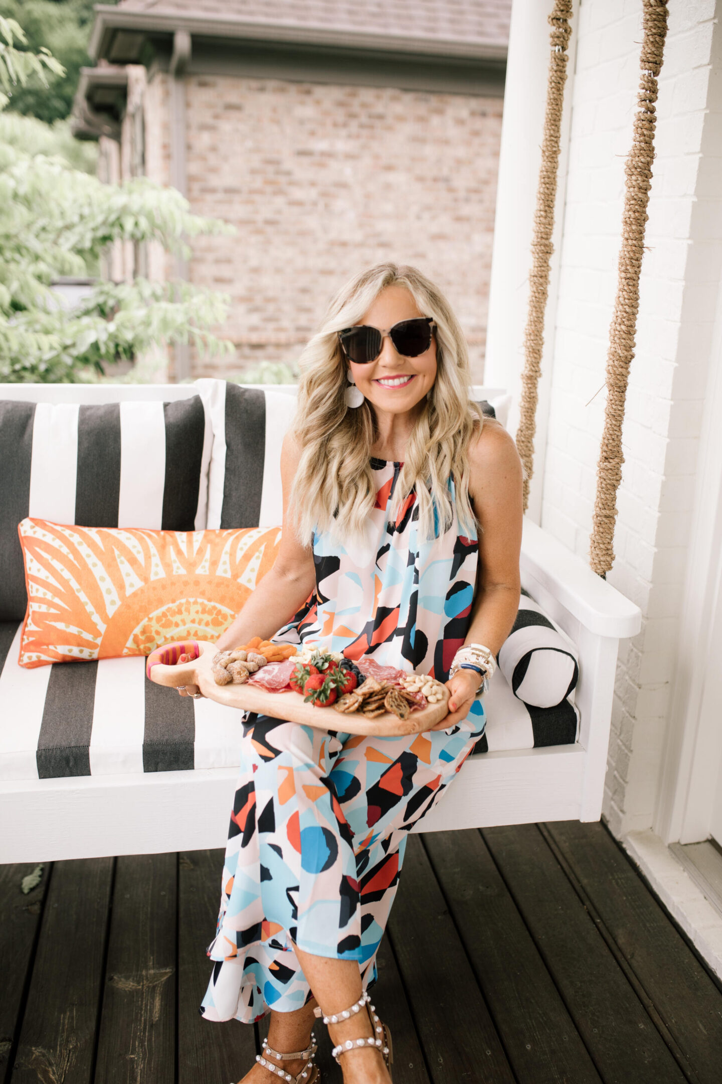 Anthropologie Products by popular Nashville life and style blog, Hello Happiness: image of Natasha Stoneking sitting on a porch swing with black and white stripe cushions and a orange and cream print lumbar pillow and wearing a geometric prints maxi dress while holding a wooden serving board filled with meat, nuts, carrot sticks and strawberries. 