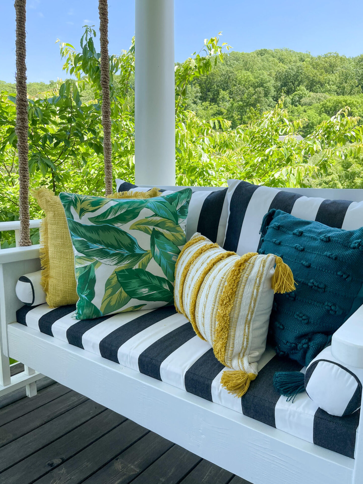 Walmart Summer by popular Nashville life and style blog, Hello Happiness: image of a porch swing with a black and white stripe cushion, yellow fringe throw pillow, palm frond print throw pillow, blue tassel pillow and yellow and white stripe lumbar pillow. 