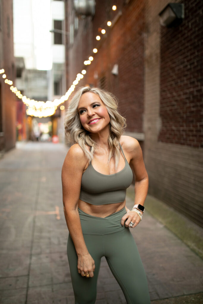 Faster Way to Fat Loss by popular Nashville lifestyle blog, Hello Happiness: image of Natasha Stoneking wearing olive green athleticwear and standing in an alley way decorated with bistro lights. 