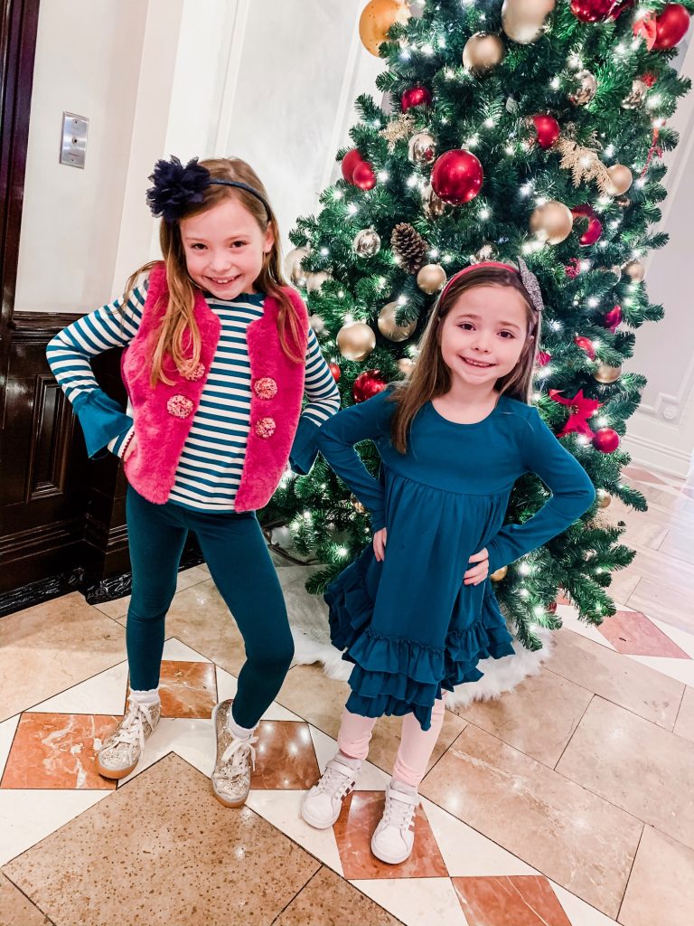 Meet Me for a Weekend in St. Louis | The Perfect Family Getaway by popular travel blog, Hello Happiness: image of two girls standing in front of a Christmas tree and wearing a pair of J.Crew Girls' everyday leggings, Matilda Jane Clothing Good to Faux Vest, Matilda Jane strip top, and Matilda Jane Clothing ruffle dress. 