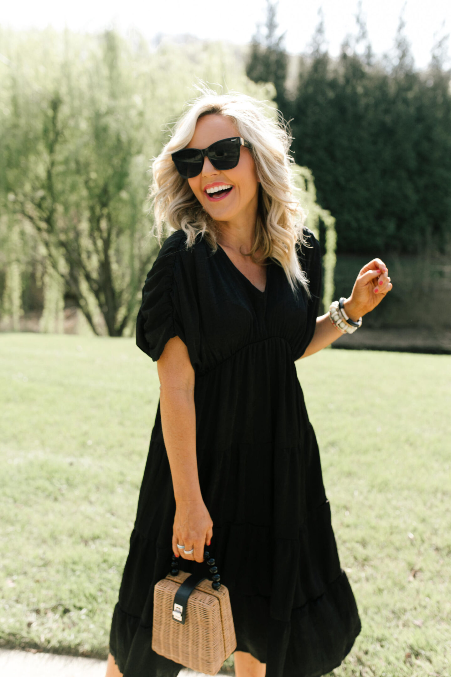 Summer Dresses by popular Nashville of Natasha Stoneking wearing a shirred tiered midi dress, after hours sunglasses, platform wedge sandals, and holding a woven bucket bag. 