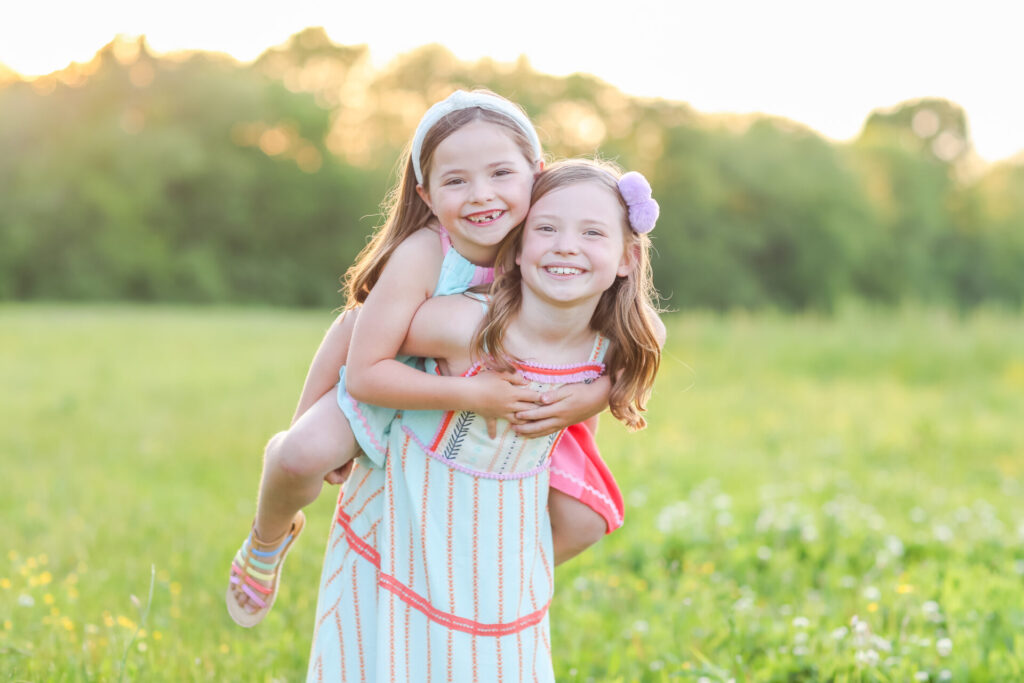Summer Bucket List by popular Nashville lifestyle blog, Hello Happiness: image of a girl giving another girl a piggy back ride in a grassy meadow and wearing Matilda Jane Clothing dresses. 