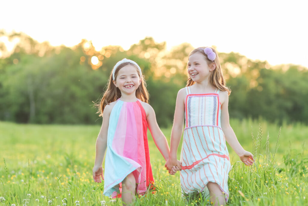 Summer Bucket List by popular Nashville lifestyle blog, Hello Happiness: image of two girls holding hands and walking through a grassy meadow and wearing Matilda Jane Clothing dresses. 